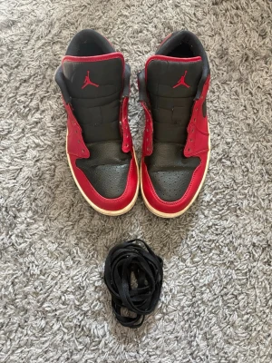 Air Jordan 1 Low svart/röd - Säljer ett par Air Jordan 1 Low i svart och rött läder med vita sulor och svarta Swoosh-loggor på sidorna. Skorna har den klassiska Jumpman-loggan på plösen och kommer med svarta skosnören. Perfekta för dig som gillar streetwear och sneakerskultur.