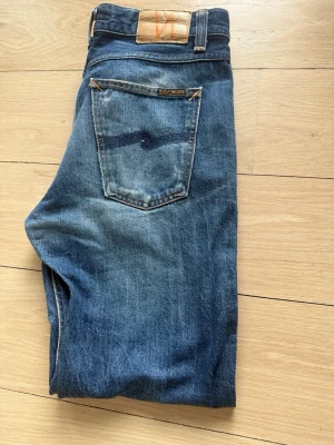 Blå jeans från Nudie Jeans - Säljer ett par klassiska blå jeans från Nudie Jeans med snygg slitning och raka ben. Jeansen har fem fickor, orange sömmar och en patch med logga bak i midjan. Tillverkade i denim av bomull. Perfekta för en avslappnad och trendig look.