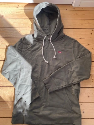 Olivgrön hoodie från Levi's - Snygg olivgrön hoodie från Levi's med klassisk huva och vita dragsnören. Liten röd Levi's-logga på bröstet och stor magficka framtill. Tillverkad i mjukt bomullsmaterial och har ribbade muddar vid ärmslut och nederkant. Perfekt för en avslappnad streetwear-look.