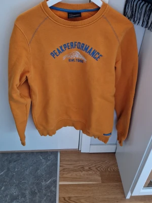 Gul sweatshirt från Peak Performance - Cool gul sweatshirt från Peak Performance med blått tryck och bergmotiv på bröstet. Rund halsringning och ribbade muddar vid ärmslut och nederkant. Perfekt för en avslappnad och sportig stil.
