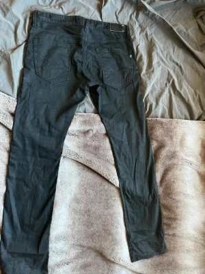 Svarta jeans från Replay - Säljer ett par svarta jeans från Replay med raka ben och klassiska bakfickor. Jeansen har en metallbricka med Replay-logga bak i midjan och en liten broderad logga på fickan. Materialet är mjukt och stretchigt för skön passform.