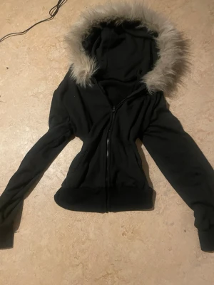 Svart hoodie med fuskpäls - Snygg svart hoodie med dragkedja och stor huva kantad med ljusgrå fuskpäls. Hoodien har långa ärmar och ribbade muddar vid ärmslut och nederkant. Perfekt för dig som vill ha en cool och mysig stil.