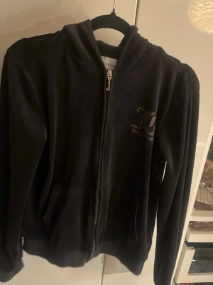 Svart Juicy Couture hoodie med dragkedja - Mjuk och skön svart hoodie från Juicy Couture i velourmaterial. Hoodien har dragkedja framtill, fickor och en liten Juicy Couture-logga på bröstet. Perfekt för en avslappnad och trendig look.
