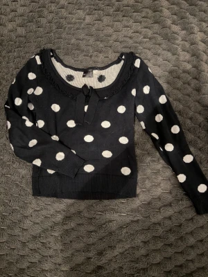 Svart stickad prickig tröja bpc - Snygg svart stickad tröja från bpc med vita stora prickar och en söt rosett vid halsringningen. Tröjan har långa ärmar, ribbade muddar och en dekorativ kant runt halsen. Perfekt för dig som gillar retrovibbar och lekfull stil.
