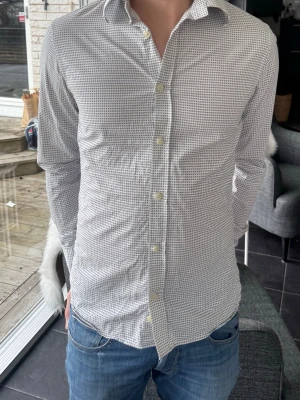 Tiger of sweden skjorta  - Snygg vit tiger of sweden  skjorta med diskret rutigt mönster och klassisk krage. Skjortan har långa ärmar och stängs med ljusa knappar framtill. Perfekt till jeans för en clean och stilren look. Det är storlek 38, 15 men passar på er som har small vanligtvis 