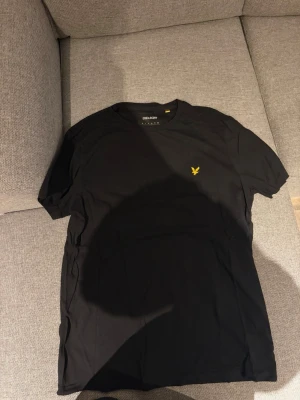 Svart t-shirt från Lyle & Scott - Klassisk svart t-shirt från Lyle & Scott med gul broderad logga på bröstet. T-shirten har rund halsringning och korta ärmar. Tillverkad i mjuk bomull för en skön känsla och enkel stil.