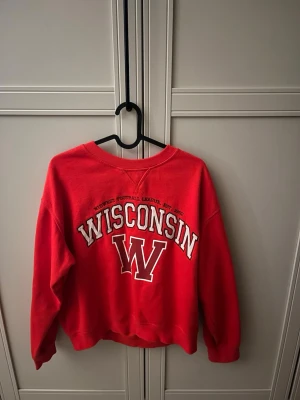Röd Wisconsin college-tröja - Cool röd sweatshirt med stort tryck 'WISCONSIN' och ett stort W på bröstet. Klassisk college-stil med rund halsringning och ribbade muddar. Perfekt för dig som gillar sportig och avslappnad look. Sweatshirt kommer från hm och är i storlek S-M