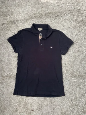 Svart pikétröja från Burberry - Klassisk svart pikétröja från Burberry med korta ärmar och den ikoniska rutiga detaljen vid knäppningen. Liten broderad logga på bröstet. Perfekt för en clean och stilren look.