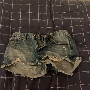 Grå slitna jeansshorts med fransar - Säljer ett par grå jeansshorts med slitna detaljer och fransig kant. Shortsen har klassisk femficksmodell, hög midja och råa avslut. Perfekta för en avslappnad och trendig look. Materialet är denim i bomull.