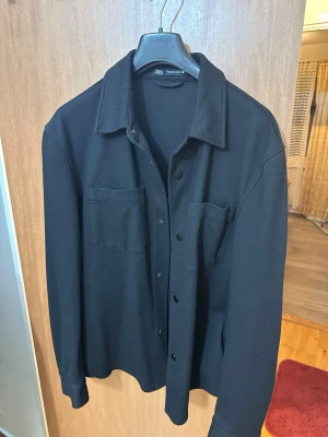 Svart overshirt från Zara - Stilren svart overshirt från Zara med klassisk krage och två bröstfickor. Skjortjackan har knäppning framtill och är tillverkad i ett mjukt, tjockare tyg som passar perfekt som lager på lager. Enkel och clean design som funkar till det mesta.