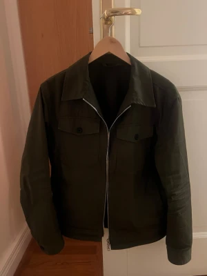 Mörkgrön overshirt med dragkedja - Stilren mörkgrön overshirt med klassisk krage och två bröstfickor med knappar. Jackan har rak passform och stängs med en silverfärgad dragkedja framtill. Materialet ser ut att vara kraftig bomull eller bomullsblandning, perfekt för lager på lager. Nypris ca 1300