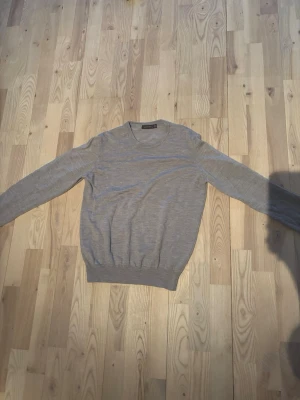 Oscar Jacobsson  - En stilren ljusgrå stickad tröja med rund halsringning och långa ärmar. Tröjan har ribbade muddar vid ärmslut och nederkant, vilket ger en klassisk och enkel look. Perfekt att matcha med jeans eller kjol.