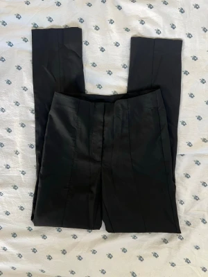 Svarta BNWT HM kostymbyxor med pressveck - Säljer ett par helt nya med lappen kvar oanvända svarta kostymbyxor från HM med raka ben och tydliga pressveck. Byxorna har hög midja och klassisk passform, perfekta till en snygg och stilren look. Materialet ser ut att vara slätt och lite glansigt, vilket ger en dressad känsla.