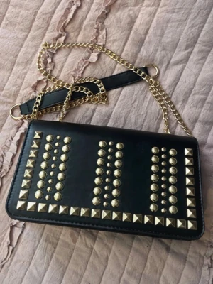Svart handväska med nitar och kedja - Cool svart handväska med guldfärgade nitar i olika former på framsidan och en lång guldfärgad kedjerem. Väskan har en rektangulär form och detaljer i metall som ger en edgy look. Perfekt accessoar för att lyfta din outfit.