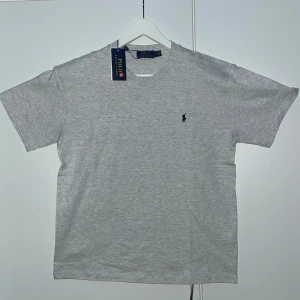 Grå t-shirt från Polo Ralph Lauren - Snygg grå t-shirt från Polo Ralph Lauren med klassisk rund hals och korta ärmar. T-shirten har en liten mörkblå broderad logga på bröstet och är gjord i mjuk bomull. Perfekt basplagg till garderoben.
