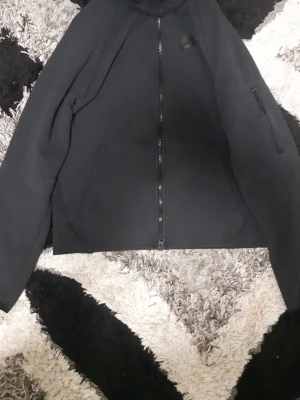 Svart Nike hoodie med dragkedja - Säljer en svart hoodie från Nike med dragkedja framtill och justerbar huva med snören. Hoodien har två stora fickor fram och en extra ficka med dragkedja på ärmen. Tillverkad i mjukt material som passar perfekt till en avslappnad stil.