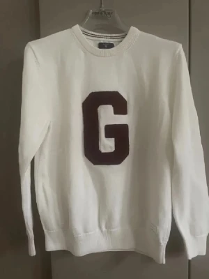 Vit stickad tröja med vinröd G - Vit stickad tröja från Gant med en stor vinröd bokstav G på bröstet. Tröjan har rund halsringning, ribbstickade muddar och är tillverkad i mjukt material. Perfekt för dig som gillar klassisk och enkel stil. Färg- gräddvit 