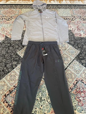 Under Armour dress - Snyggt träningsset från Under Armour med en ljusgrå hoodie med dragkedja och huva samt matchande mörkgrå byxor med resårmidja och dragsko. Setet har logga på både jacka och byxa samt sidofickor. Perfekt för träning eller chill. Materialet är lätt och andas.