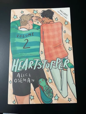 Heartstopper  2  - Heartstopper Volym 2 är en grafisk roman med färgstarkt illustrerat omslag. Boken handlar om kärlek och vänskap mellan två tonårskillar. Omslaget har tecknade figurer i gröna och orange toner och boken är tryckt på papper.