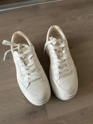 Vita chunky sneakers med platåsula - Säljer ett par helvita sneakers med chunky platåsula och breda snören. Skorna har en stilren design och är tillverkade i syntetmaterial med textildetaljer. Perfekta för dig som gillar minimalistisk och trendig stil.