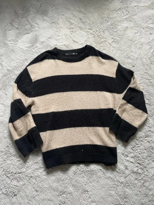 Beige och svart randig stickad tröja - Stickad tröja från 157 i beige och svart med breda horisontella ränder. Tröjan har rund halsringning och långa ärmar med ribbstickade muddar. Materialet är mjukt och lite fluffigt, perfekt för att hålla sig varm under kyliga dagar.