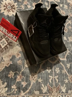Svarta Air Jordan 4 Retro sneakers - Säljer ett par helsvarta Air Jordan 4 Retro sneakers med klassisk meshpanel och läderdetaljer. Skorna har högt skaft, ikonisk Jumpman-logga på plösen och coola plastdetaljer vid snörningen. Perfekta för dig som gillar streetwear och vill sticka ut. Dom är lite använda 