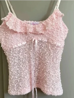 Ljusrosa linnetopp med volang från Loana - Supersöt ljusrosa linnetopp från Loana med tunna axelband, volang vid urringningen och dekorativt band med rosett framtill. Toppen har ett fluffigt, stickat material som ger en unik och gullig look. Perfekt för sommarens alla tillfällen! Köpt i Frankrike 