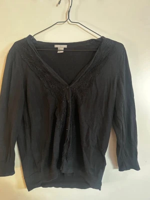 Svart kofta med spetsdetaljer - Svart kofta från H&M med knäppning framtill och fina spetsdetaljer längs den djupa V-ringningen. Koftan har trekvartsärmar och är tillverkad i ett mjukt, tunt material som passar perfekt till lager på lager-stil. Rök och djurfritt hem. Pruta gärna 🌸