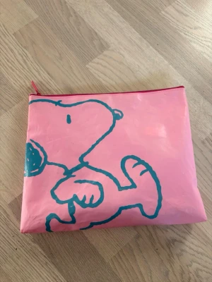 Rosa necessär med Snoopy-tryck - Söt rosa necessär med ett stort turkost Snoopy-tryck på framsidan. Väskan har dragkedja upptill och är tillverkad i ett plastliknande material. Perfekt för småprylar eller smink. Enkel och lekfull design som sticker ut. Använd se bilder men dragkedjan funkar bra 