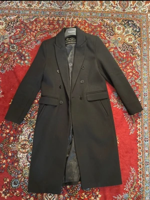 Svart Amisu Kappa - Stilren svart lång kappa. Kappan har dubbelknäppning, två fickor med lock och klassisk krage. Fodrad insida för extra komfort och en tidlös look som passar till många olika stilar. Köpt från New yorker för ca 600 kr. Den är dock lite nopprig, och en knapp är lite lös, som visas i sista bilden. 