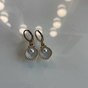 Guldpläterade snäckformade örhängen - Säljer ett par örhängen i snäckform med guldfärgad metall och en vit pärldetalj i mitten. Örhängena har en klassisk klickstängning och en elegant, havsinspirerad design. Perfekta för dig som gillar unika accessoarer med en touch av strandkänsla.