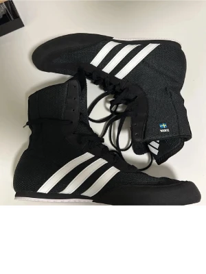 Svarta boxningsskor från Adidas - Säljer ett par svarta boxningsskor från Adidas med klassiska vita ränder på sidorna. Skorna har hög modell med snörning hela vägen upp och tunn, flexibel sula. Materialet är lätt och andas, perfekt för träning och match. Svensk flagga och texten 'VNZ' på sidan.