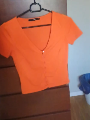 Orange croppad topp med knappar - Snygg orange croppad topp från Bik Bok med korta ärmar och v-ringning. Toppen har knappar framtill och är gjord i ett mjukt, stretchigt material som sitter tight. Perfekt för dig som vill ha en färgstark och trendig look.