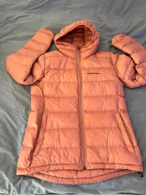 Rosa dunjacka från Peak Performance - Säljer en snygg rosa dunjacka från Peak Performance med huva och dragkedja framtill. Jackan har två sidofickor med dragkedja och quiltad design som ger extra värme. Perfekt för kalla vinterdagar och riktigt skön att ha på sig.