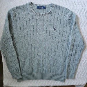 Ljusgrå kabelstickad tröja Polo Ralph Lauren - Snygg ljusgrå kabelstickad tröja från Polo Ralph Lauren med rund halsringning och klassisk broderad logga på bröstet. Tröjan har ribbade muddar vid ärmslut och nederkant. Perfekt för lager på lager och en stilren look.