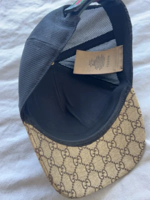 Gucci keps med tigertryck och GG-mönster - Snygg keps från Gucci med klassiskt beige GG-mönster och två stora tigertryck framtill. Baksidan är i svart mesh för extra ventilation och justerbar passform. Perfekt för dig som vill sticka ut med en exklusiv accessoar.                           Ingen box tyvärr