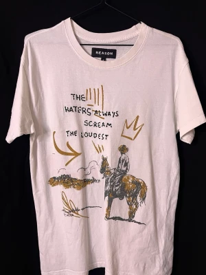 Vit t-shirt med tryck från Reason - Vit t-shirt från Reason med coolt tryck framtill som visar en cowboy på häst och texten 'The haters always scream the loudest' i svart och guld. T-shirten har rund hals och korta ärmar. Perfekt för dig som gillar streetwear och unika prints. Skick 8/10 med ett litet hål på baksidan.