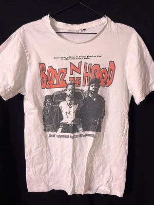 Vit Boyz N The Hood t-shirt - Säljer en vit t-shirt med stort Boyz N The Hood-tryck fram och bak. Framsidan har ett ikoniskt filmomslag i svartvitt med röd text och detaljer, medan baksidan har loggan och texten 'Increase the Peace'. Klassisk passform och rund hals. Perfekt för dig som gillar streetwear och filmreferenser.