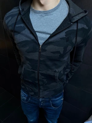 Polo ralph lauren hoodie - Snygg svart hoodie från Polo ralph lauren med mörkgrått camomönster och dragkedja framtill. Modellen har huva med dragsko och två fickor på magen. Perfekt för en avslappnad stil. Materialet känns mjukt och bekvämt mot huden.