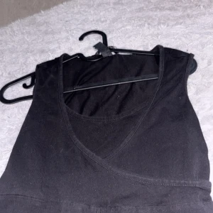 Svart omlott linnetopp - Snygg svart ärmlös linnetopp med omlottdetalj framtill. Perfekt att matcha med jeans eller kjol för en trendig look. Toppens material känns mjukt och följsamt mot kroppen och den har en djup v-ringning.