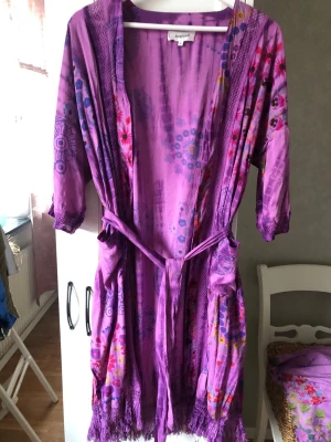 Lila mönstrad tunika Desigual Oanvänd - Säljer en helt oanvänd  lila tunika från Desigual med färgglada blommönster och grafiska detaljer. Tunikan har trekvartsärmar, knytskärp i midjan och dekorativa fransar i nederkant. Den har 2 fickor.  Se även matchande byxor i annan annons!