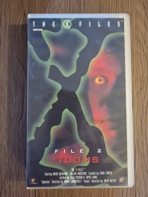 The X-Files File 2 Tooms VHS - Säljer en VHS-kassett med The X-Files: File 2 Tooms. Omslaget har en stor grön X och ett ansikte i bakgrunden. Text på svenska på baksidan och klassisk plastförpackning. Perfekt för samlare av retrofilm och sci-fi.