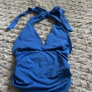 Blå halterneck topp - Snygg blå topp med halterneck och knytband i nacken. Modellen har djup urringning och rynkade detaljer i sidorna för en schysst passform. Tillverkad i stretchigt material som känns bekvämt på stranden eller vid poolen.