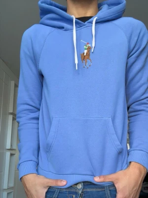 Blå hoodie från Polo Ralph Lauren - Snygg blå hoodie från Polo Ralph Lauren med klassisk broderad polospelare på bröstet. Hoodien har vit dragsko, känguruficka och ribbade muddar. Tillverkad i mjukt bomullsmaterial som är skönt mot huden.