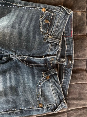 True Religion jeans - Snygga low Rise flare jeans från True religion med coola nitar längs fickor och bensidor. Jeansen har klassisk femficksmodell, dragkedja och knapp i midjan samt slitningar för en trendig look. Tillverkade i stretchigt denim för skön passform.