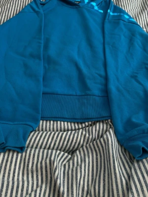 Blå hoodie med huva - Snygg blå hoodie med huva och ribbade muddar vid ärmslut och nederkant. Hoodien har en klassisk passform och är tillverkad i mjukt material som känns skönt mot huden. Perfekt för en avslappnad och cool stil.