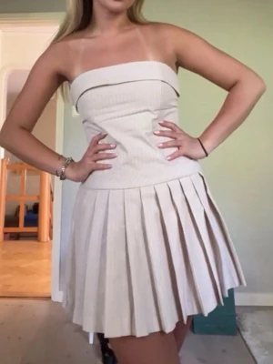 Beige axelbandslös kort klänning - Säljer en beige/rosa axelbandslös kort klänning med tunna vita ränder och plisserad kjol. Klänningen har en söt volangdetalj upptill och är perfekt för sommarens alla tillfällen.