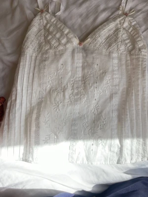 Vitt spetsnattlinne med broderi - Romantiskt vitt nattlinne med tunna axelband, spetsdetaljer över bysten och vackra broderier framtill. Små rosa rosetter vid axelbanden och dekorativa vertikala spetsband. Luftigt och mjukt material, perfekt för mysiga kvällar.