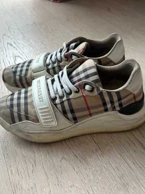Burberry rutig sneakers med snörning - Säljer ett par klassiska Burberry sneakers med det ikoniska rutiga mönstret i beige, svart och rött. Skorna har vita detaljer, snörning och ett brett kardborreband med Burberry-logga. Snygg chunky sula och bekväm passform. Perfekta för dig som gillar statement-plagg.