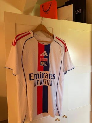 Olympique Lyonnais matchtröja Adidas - Säljer en vit Olympique Lyonnais matchtröja från Adidas med röda och blå detaljer. Tröjan har klubbmärke och Adidas-logga på bröstet, samt sponsortryck 'Emirates Fly Better' framtill. Klassiska röda ränder på axlarna och korta ärmar. Perfekt för fotbollsfans!